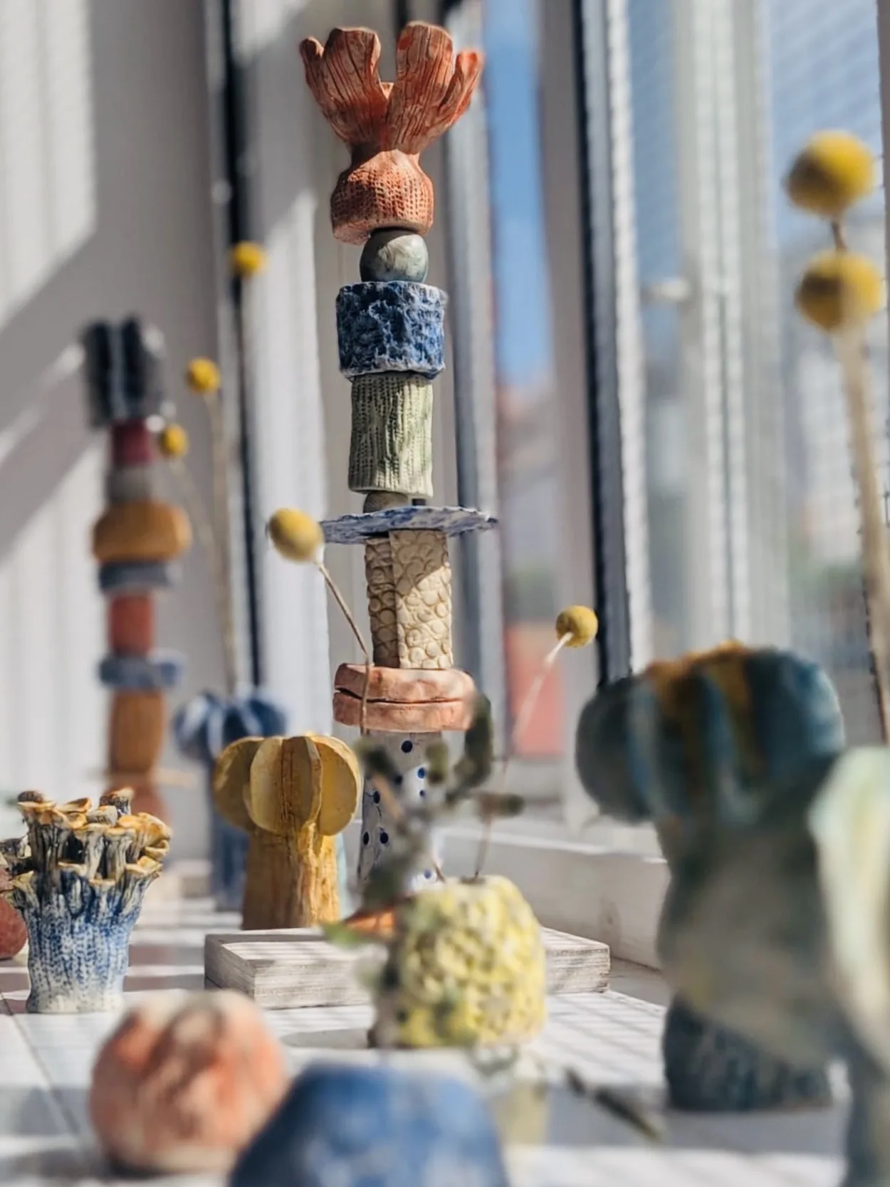 Textured, colourful stacked ceramic totems and pots lined up along a sunny windowsill